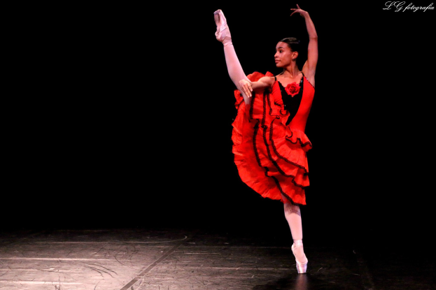 Eine Frau in einem roten Kleid tanzt auf der Bühne Ballett, Arme und Beine ausgestreckt, trägt Pointe-Schuhe und einen ordentlichen Dutt.