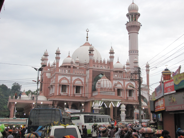 Eine große Menschenmenge geht eine Straße entlang vor einer Moschee, mit Fahrzeugen und Fußgängern, einige halten Schirme, unter einem klaren blauen Himmel.