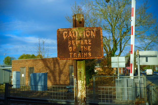 Vorsichtsschild an einem Bahnübergang-Zaun mit Bäumen, Strommasten, einem Gebäude, Containern, Fahrzeugen auf einer Straße und einem bewölkten Himmel.