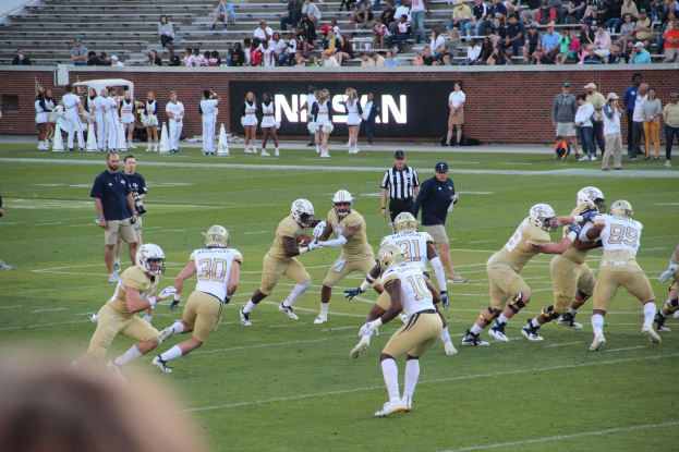 Menschen in Sportbekleidung und Helmen spielen Fußball auf einem Feld, während andere Kameras halten, mit Zuschauern auf Bänken und einer textbedeckten Wand im Hintergrund.