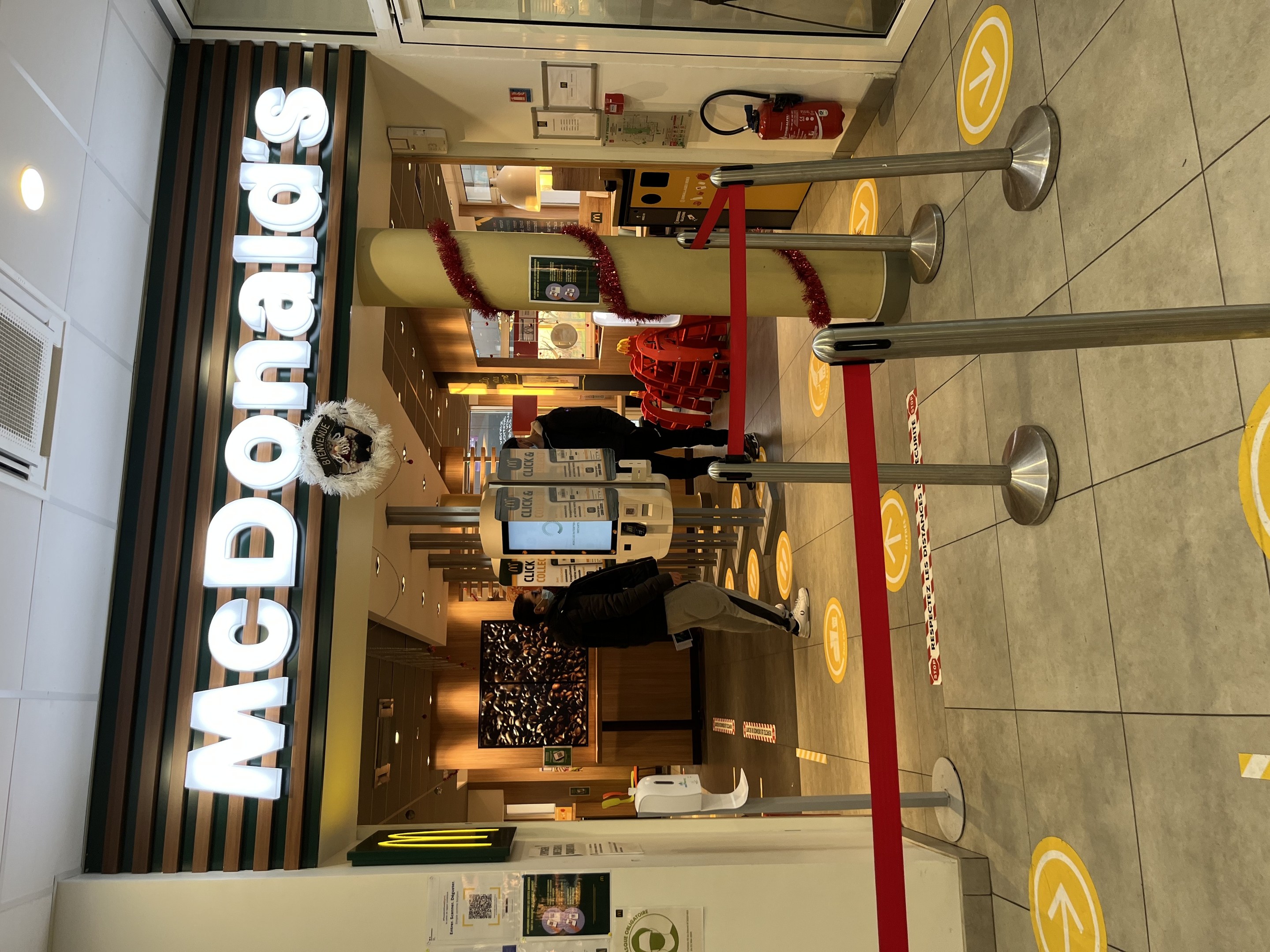 Innenansicht eines McDonald's-Restaurants mit einem sichtbaren Schild, Metallstangen mit roten Bändern und einigen Menschen, die davor stehen.