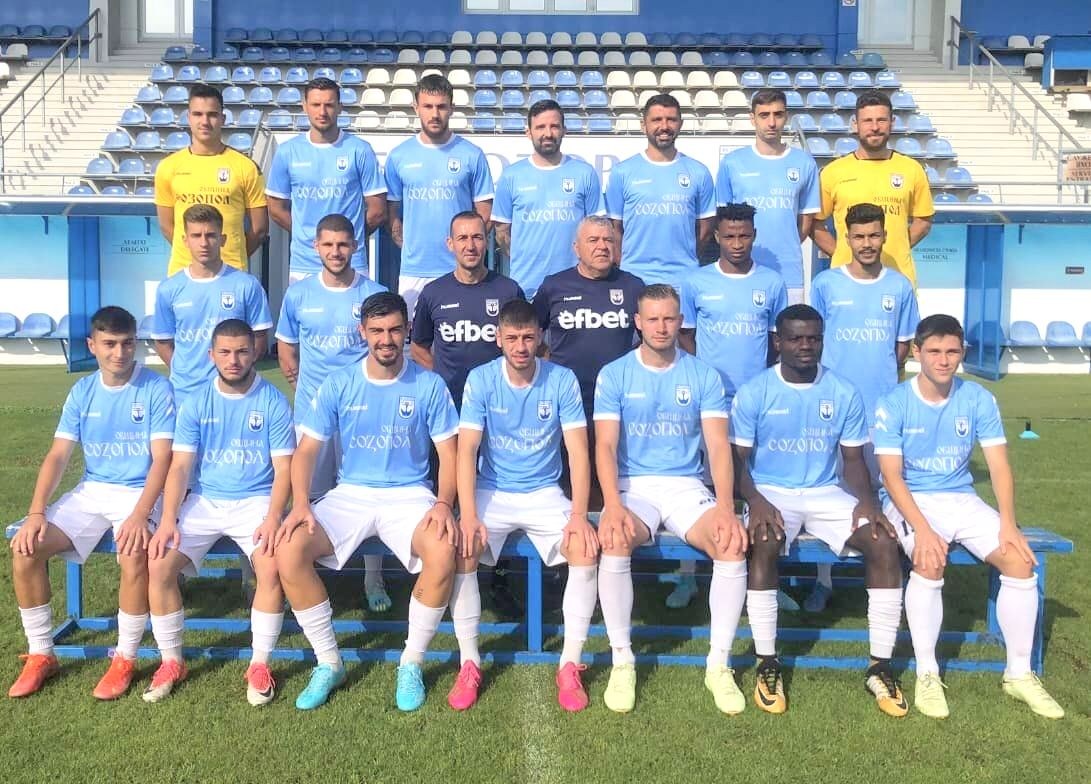 Eine Gruppe von Männern, einige sitzen auf Bänken und andere stehen auf einem Fußballfeld, mit einem Stadion-Sitzbereich und architektonischen Elementen im Hintergrund, was einen siegreichen Team nach einem Spiel vermuten lässt.