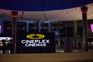 Das Innere des Cineplex Cinemas bei Nacht, beleuchtet von Deckenlampen, mit Säulen, Texttafeln, Geländern, Glaswänden, Bildschirmen und wenigen Menschen, die eine dunkle und mysteriöse Atmosphäre schaffen.