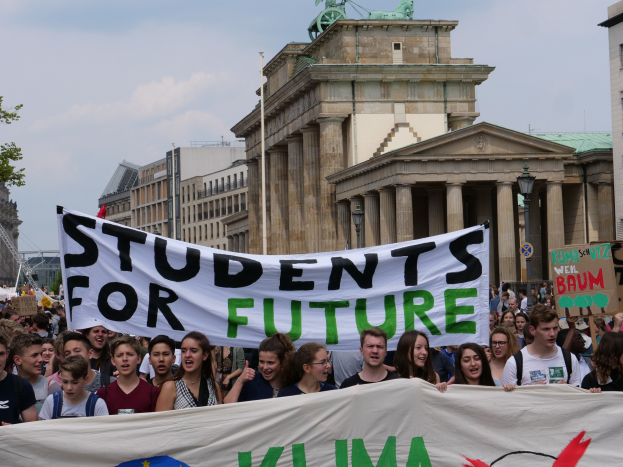 Eine Gruppe von Schülern marschiert in Berlin mit einem bunt bemalten "Students for Future"-Schild an Gebäuden, Bäumen und Himmel vorbei.