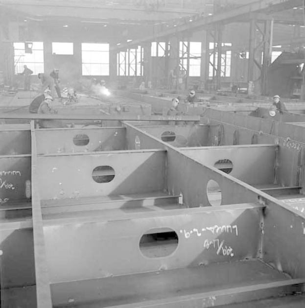 Ein Schwarz-Weiß-Foto einer Fabrik mit Arbeitern in Helmen, die an Stahlträgern arbeiten, mit Fenstern, Pfählen, Lichtern und Text im Hintergrund.