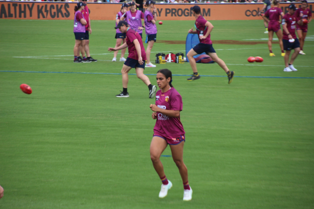 Frauen spielen Australian Rules Football auf einem Rasenfeld mit verstreuten Bällen und einem Banner im Hintergrund, einige tragen Kappen und Turnschuhe.