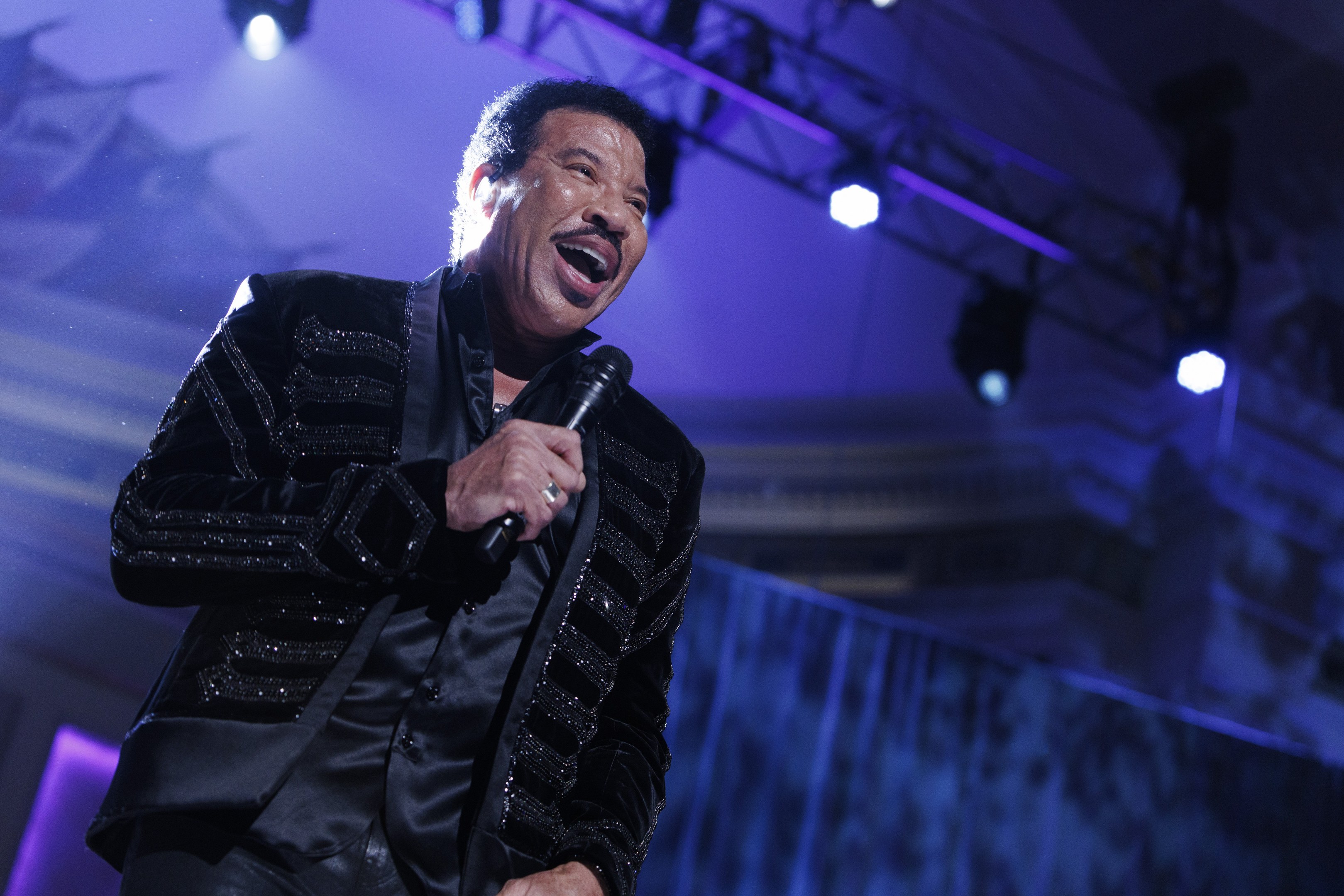 Lionel Richie performs bei den Grammys, tr├Ągt ein schwarzes Kleid und h├Ąlt ein Mikrofon auf einer hell erleuchteten Bühne.