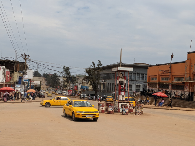 Ein gelbes Taxi fährt eine belebte Stadtstraße mit Gebäuden an den Seiten hinunter, während Fußgänger mit Schirmen auf dem Gehweg darunter unter bewölktem Himmel und Stromleitungen oben gehen.