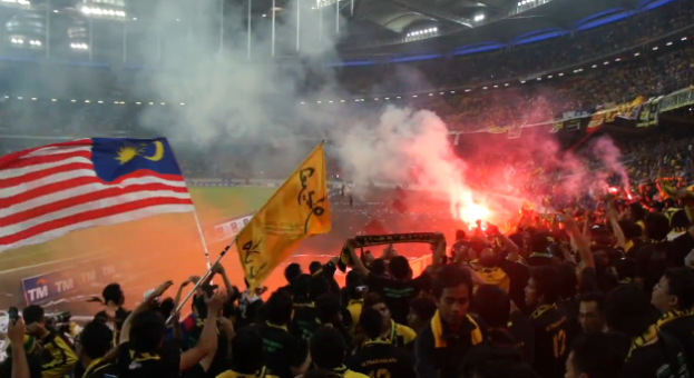 Eine große Menschenmenge in einem Stadion mit Bengalos, Fahnen und einer Kamera, mit Rauch und Schildern mit der Aufschrift "Malaysia vs Malaysia Live-Streaming" im Hintergrund.