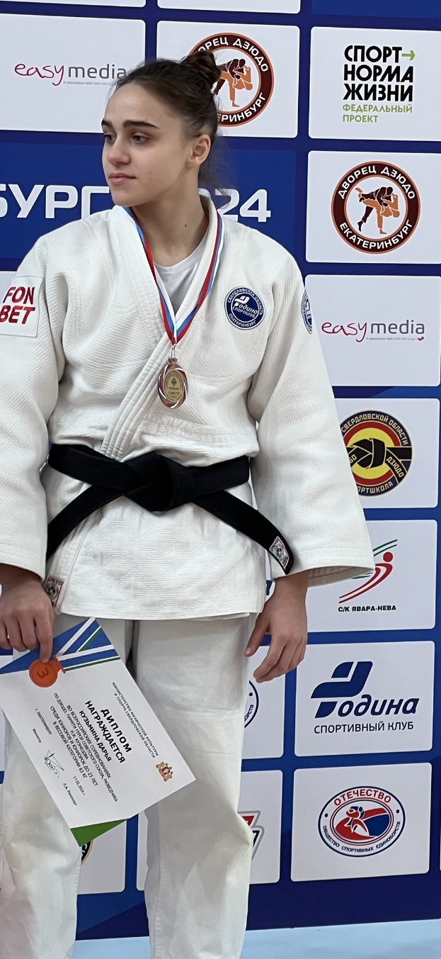Frau in weißem Judo-Anzug mit schwarzem Gürtel auf einem Podest stehend, eine Medaille und ein Zertifikat haltend, mit einem Logo-Plakat im Hintergrund.