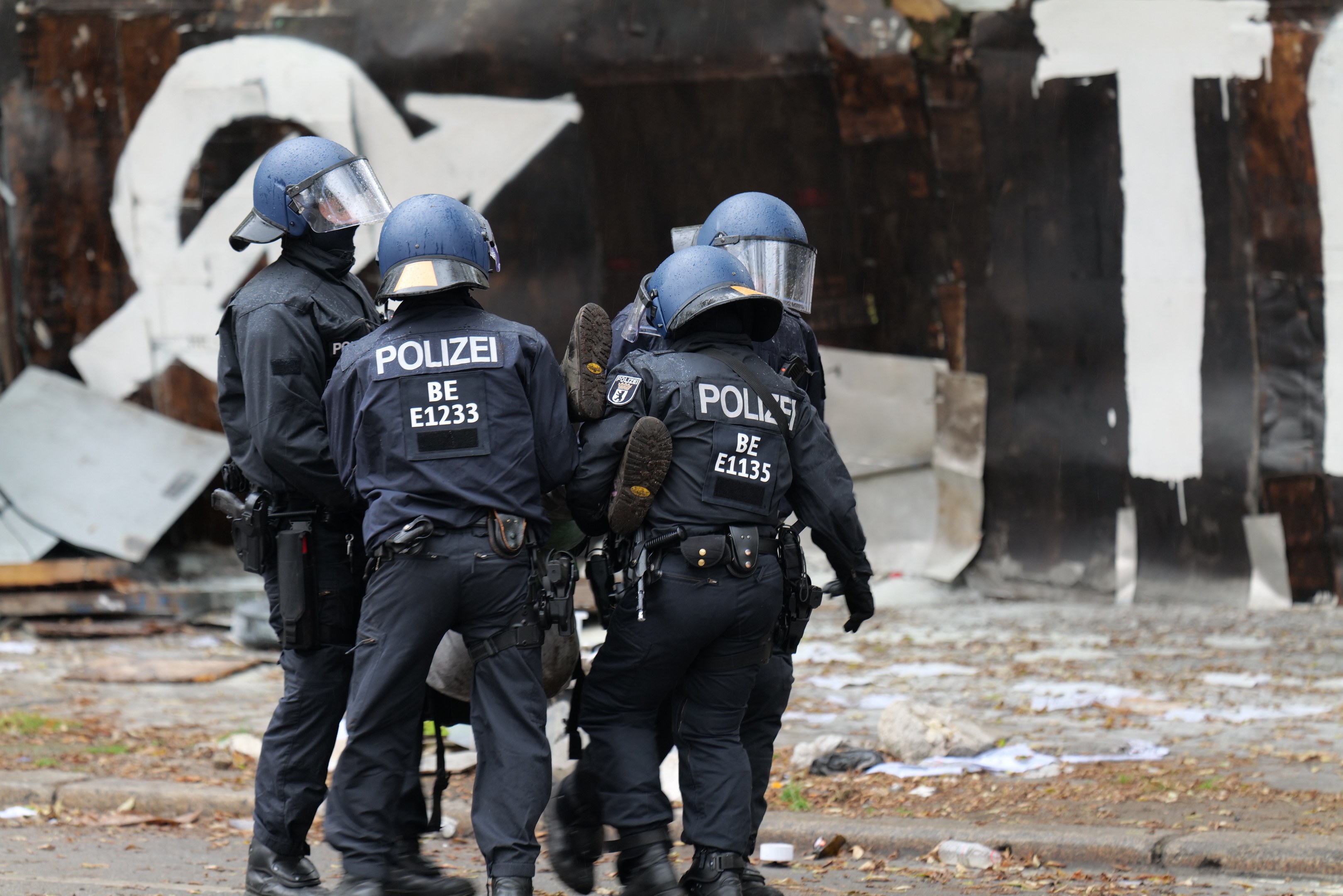 Polizeibeamte in schwarzen Uniformen und blauen Helmen stehen vor einem Gebäude mit Graffiti während einer Unruhen, mit verstreuten Papieren und Gegenständen auf dem Boden.