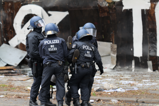 Polizeibeamte in schwarzen Uniformen und blauen Helmen stehen vor einem Gebäude mit Graffiti während einer Unruhen, mit verstreuten Papieren und Gegenständen auf dem Boden.
