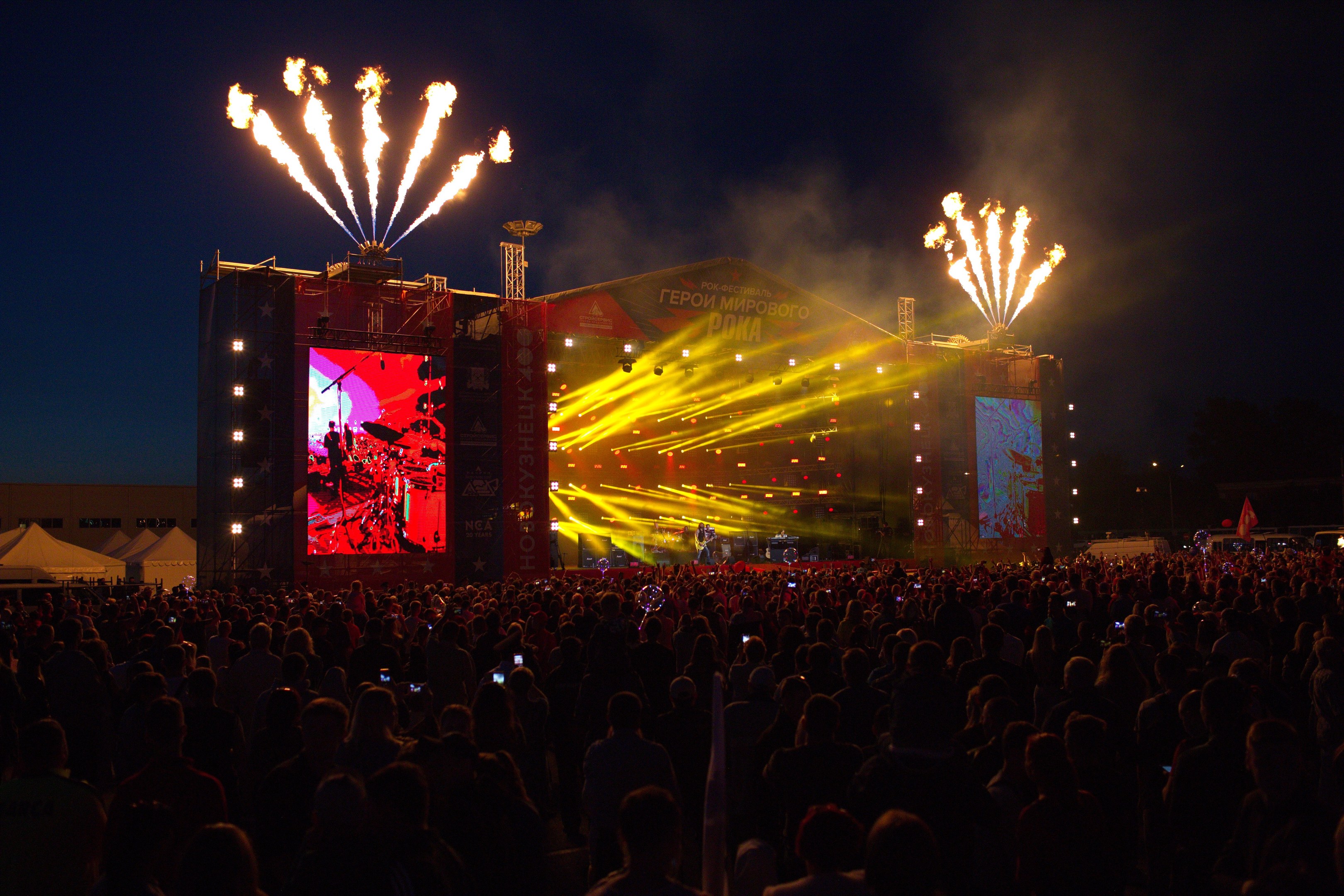 Eine große Menschenmenge bei einem Musikfestival schaut auf einer hell erleuchteten Bühne mit Bannern, Zelten links und Feuerwerk am dunklen Himmel.