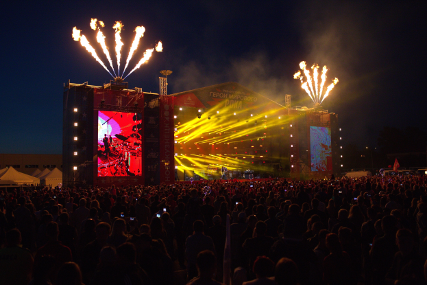 Eine große Menschenmenge bei einem Musikfestival schaut auf einer hell erleuchteten Bühne mit Bannern, Zelten links und Feuerwerk am dunklen Himmel.