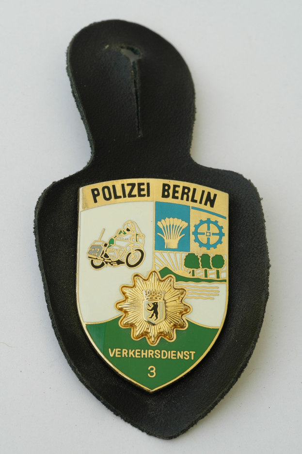 Ein schildförmiges Polizeiabzeichen mit "Polizei Berlin" in schwarzer fetter Schrift auf einem blauen Hintergrund mit einem weißen Stern und Rahmen, das auf einer Oberfläche ruht.
