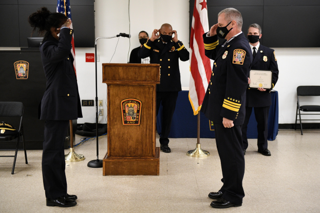 Gruppe uniformierter Personen in Masken vor einem Rednerpult mit Mikrofon, umgeben von Fahnen, Stühlen, Tischen und einer Wand während einer Vereidigung eines neuen Feuerwehrchefs, wobei eine Person ein Zertifikat hält.