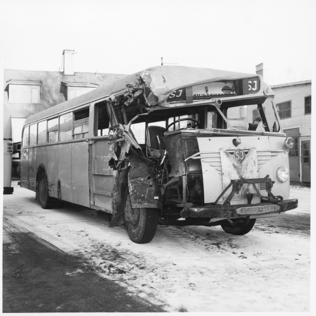 Schwarz-weißes Foto eines schwerbeschädigten Busses am Straßenrand, mit eingedellter Front und zersplitterten Fenstern, Gebäude im Hintergrund.