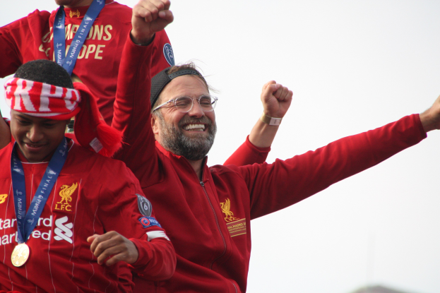 Liverpool-Trainer Jürgen Klopp feiert mit Spielern und Stab nach dem Gewinn der UEFA Champions League, trägt eine rote Jacke mit zwei medalletragenden Personen neben ihm vor einem Himmel-Hintergrund.