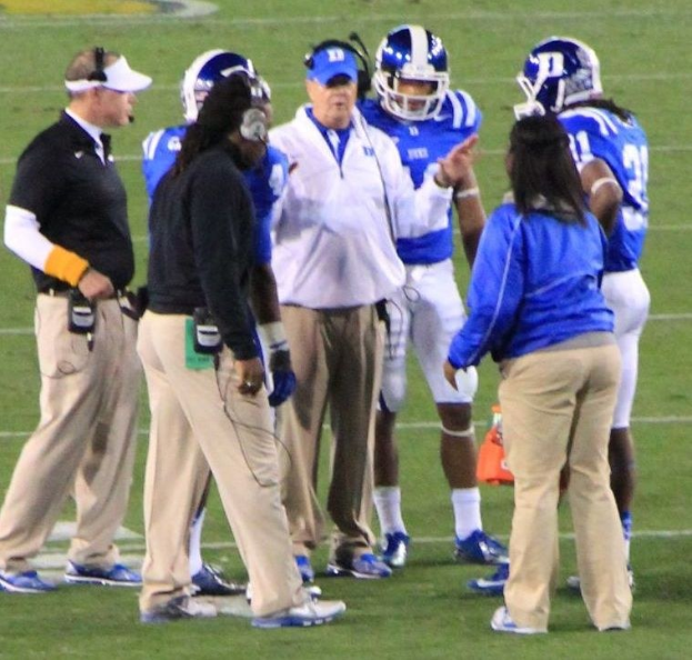 Gruppe von Menschen in Diskussion auf einem grünen Fußballfeld, einige tragen Kappen und Helme, mit einer Wand im Hintergrund.