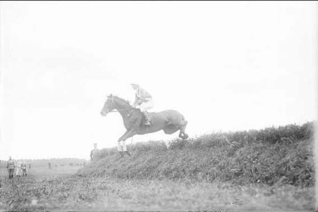 Schwarzes und weißes Foto eines Pferdes und Reiters, die über einen grasbewachsenen Hügel springen, während das Pferd galoppiert und der Reiter einen Helm und einen Mantel trägt, während Zuschauer im Hintergrund zuschauen.