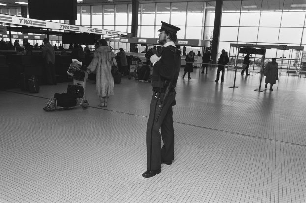 Polizeibeamter in Uniform mit Waffe in einem Flughafen-Terminal umgeben von Menschen und Gepäck, mit Gepäckwagen, Deckenleuchten, Glaswänden und Informationsanzeigen im Hintergrund.