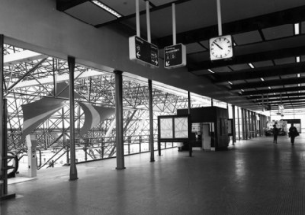 Ein Schwarz-Weiß-Foto eines Flughafen-Terminals mit Menschen, Pfählen, Brettern, einer Uhr und Deckenleuchten.