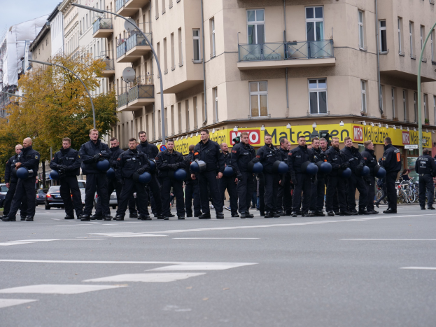 Eine Gruppe von Polizisten in schwarzen Uniformen mit blauen Helmen steht in einer Straße mit Laternen, Bäumen und Glasfenstern unter einem klaren blauen Himmel.