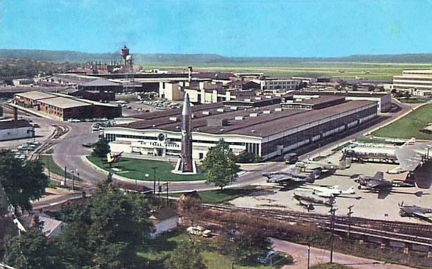 Altes Schwarz-Weiß-Foto eines Flughafens mit geparkten Flugzeugen, Gebäuden, Bäumen, Straßen, Fahrzeugen, Strommasten, Gras, Bahnschienen, Türmen, Hügeln und einem klaren blauen Himmel, mit Text unten.