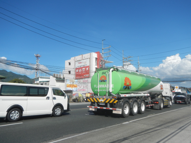 Großer Tanklastzug fährt auf einer Straße neben einem weißen Van mit Strommasten, Gebäuden, Bäumen, Hügeln und einem bewölkten Himmel im Hintergrund.