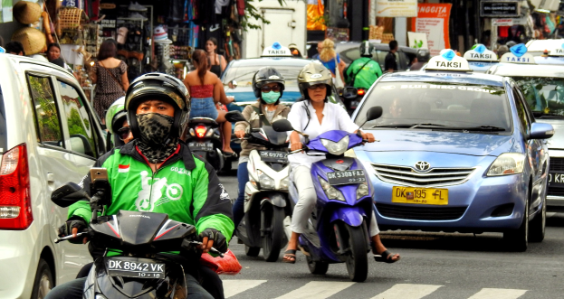 Eine Gruppe von Menschen, die Motorräder auf einer belebten Straße fahren, mit Autos und anderen Fahrzeugen, einige tragen Helme, vor einem Hintergrund von Gebäuden, Bäumen, Schildern und Ladengeschäften.