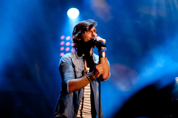 Ein Mann steht auf einer hell erleuchteten Bühne und singt in ein Mikrofon, während er ein schwarzes T-Shirt, Jeans und eine Gitarre trägt und ein selbstbewusstes Aussehen und wilde Haare hat.