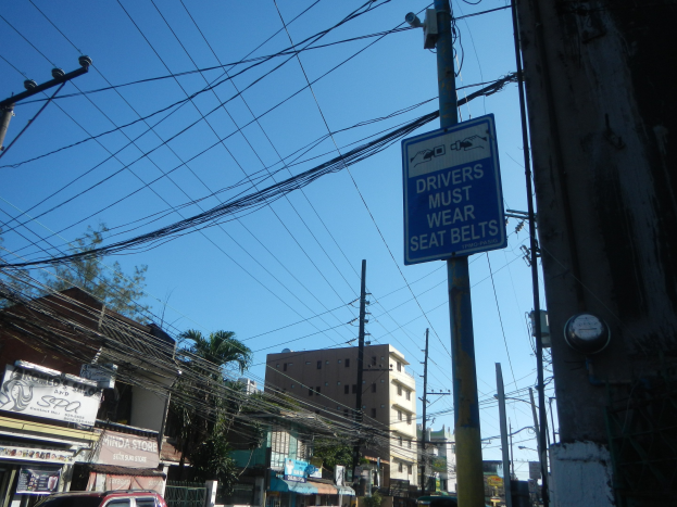 Stadtstraße mit fahrenden Autos, Strommasten mit Drähten, Gebäude mit Fenstern, Bäume, Namenschilder und ein "Fahrer müssen Sicherheitsgurte tragen"-Schild an einem Strommasten vor einem sichtbaren Himmel.