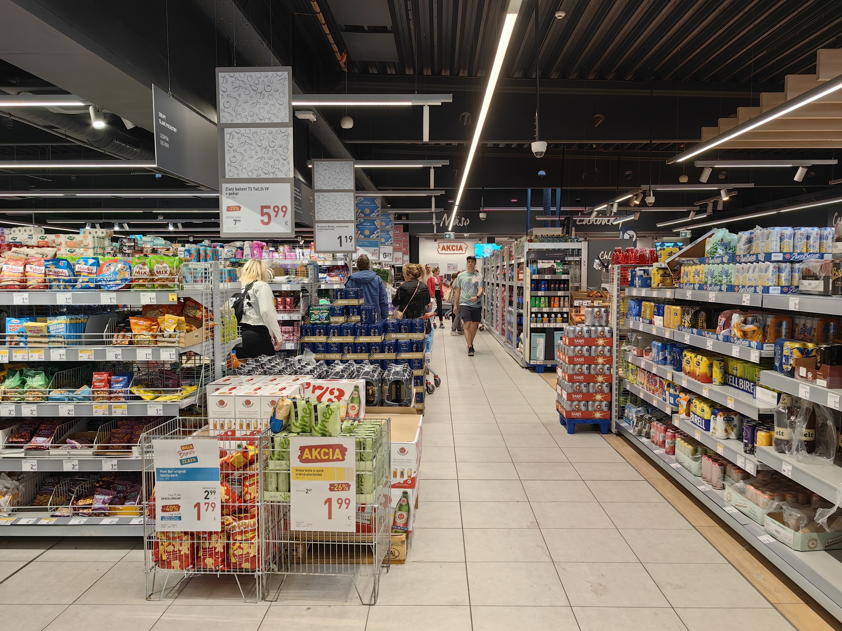 Innenansicht eines Supermarkts mit Einkäufern, Lebensmittelregalen, Einkaufswagen, Preistafeln, Deckenleuchten und sichtbarem Boden.