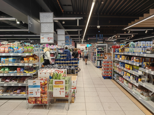Innenansicht eines Supermarkts mit Einkäufern, Lebensmittelregalen, Einkaufswagen, Preistafeln, Deckenleuchten und sichtbarem Boden.