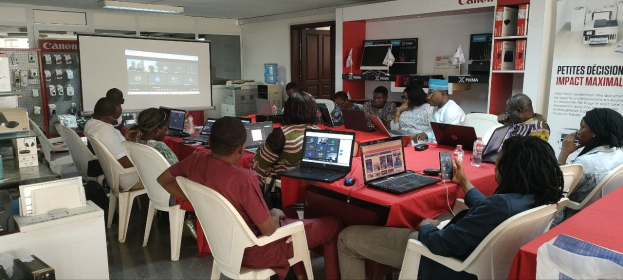 Gruppe von Menschen an einem Tisch mit Laptops und anderen Gegenständen während eines digitalen Marketing-Workshops, mit einem Banner, einem Bildschirm und Deckenleuchten im Raum.