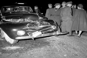 Ein Schwarzweiss-Bild eines auf der Strasse liegenden Wagens, umgeben von einer Gruppe von Menschen, die die erheblichen Schäden am Fahrzeug begutachten.