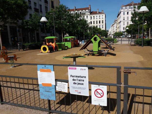 Spielplatz auf einem Stadtplatz umgeben von einem Zaun mit Schildern, Bäumen, Laternenmasten und Gebäuden im Hintergrund unter einem sichtbaren Himmel.