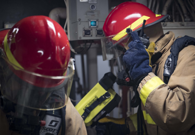 Zwei Feuerwehrleute in Schutzausrüstung arbeiten an einem Hydranten während einer Übung mit Maschinen und Kabeln im Hintergrund.