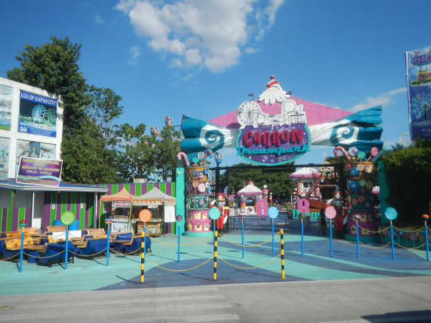 Karussell am Eingang eines Parks mit Straße, Umzäunung, Buden, Schildern, Bäumen und bewölktem Himmel im Hintergrund.