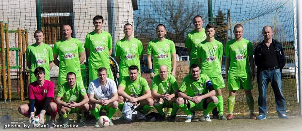 Eine Gruppe von Männern in grünen T-Shirts steht und hockt auf einem Fußballfeld mit einem Ball davor, ein Torpfosten rechts und ein Hintergrund aus Bäumen, Gebäuden, Pfählen, Fahrzeugen und dem Himmel, mit Text in der linken unteren Ecke, der die Nummern von 1 bis 72 auflistet.
