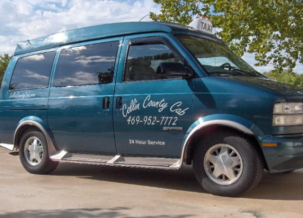 Ein blauer Lieferwagen mit Text und Zahlen darauf, der auf einem von Bäumen umgebenen Parkplatz unter einem klaren Himmel geparkt ist, wahrscheinlich ein Taxi.