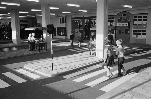 Schwarz-weiß-Foto von Kindern, die eine Straße überqueren, mit Menschen auf Bänken, einer Ampel und Hintergrundsäulen mit Deckenleuchten.