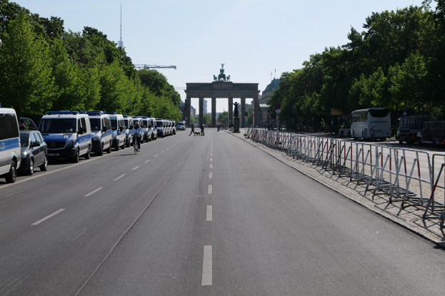 Lange Reihe von Polizeiwagen vor dem Brandenburger Tor geparkt, mit Fahrradfahrern und Passanten, Barrieren, Bäumen und einem Bogen mit Statuen im Hintergrund.