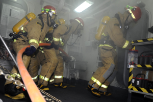 Feuerwehrleute in Schutzausrüstung mit Helmen und Sauerstoffflaschen arbeiten an einem Löschfahrzeug, mit einem Gestell voller Gegenstände und Hintergrundausrüstung im Hintergrund.
