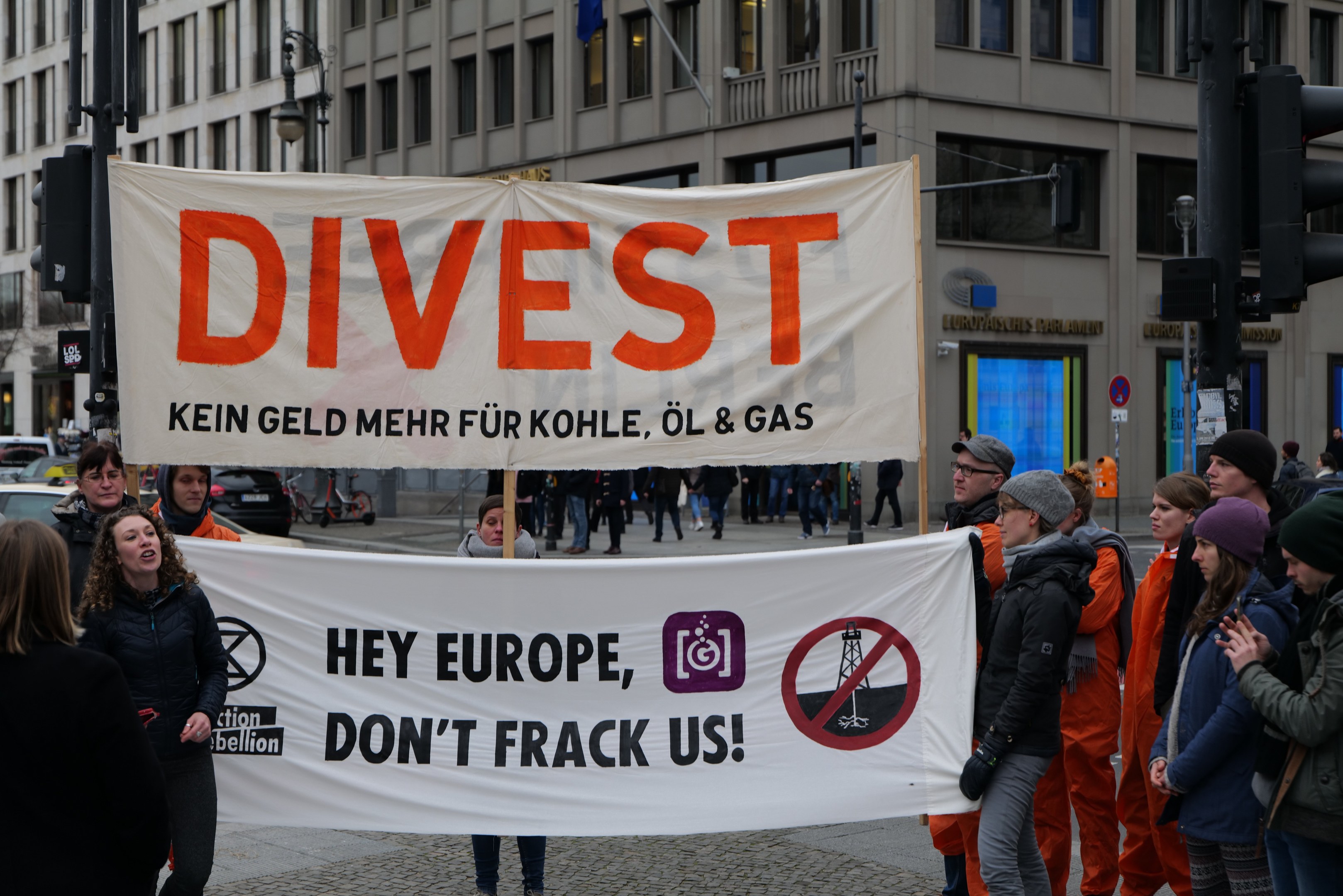 Gruppe von Menschen mit einem "Divest Hey Europe, Don't Frack Us"-Schild auf einer Straße mit Gebäuden, Straßeninfrastruktur, Fahrzeugen, Bäumen und einem klaren Himmel im Hintergrund.