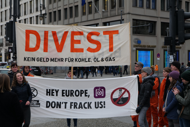 Gruppe von Menschen mit einem "Divest Hey Europe, Don't Frack Us"-Schild auf einer Straße mit Gebäuden, Straßeninfrastruktur, Fahrzeugen, Bäumen und einem klaren Himmel im Hintergrund.