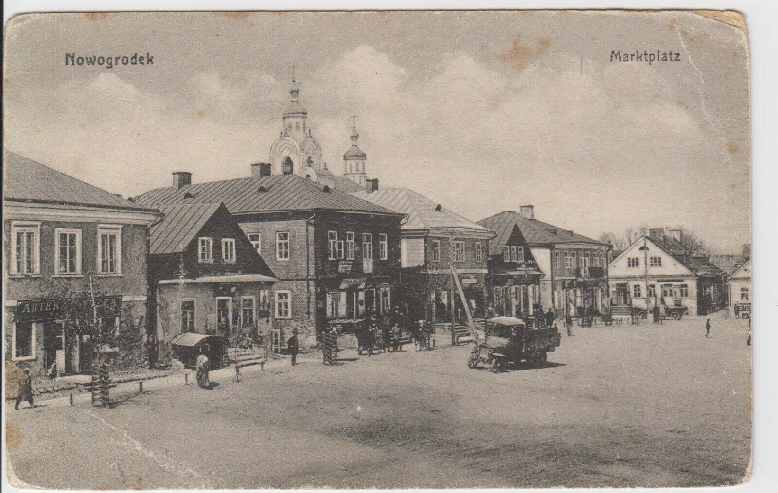 Schwarz-weiß-Fotografie einer Stadtstraße in Nowogrodeek, Deutschland, mit Gebäuden, Menschen, Karren, Pfählen, Bäumen und Himmel sowie dem Text "Marktplatz in Nowogrodeek, Deutschland" oben und unten.