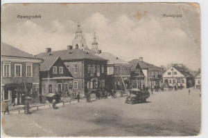 Schwarz-weiß-Fotografie einer Stadtstraße in Nowogrodeek, Deutschland, mit Gebäuden, Menschen, Karren, Pfählen, Bäumen und Himmel sowie dem Text "Marktplatz in Nowogrodeek, Deutschland" oben und unten.