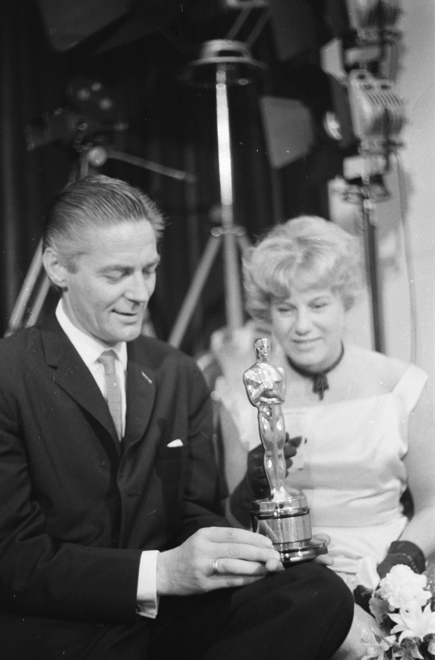 Ein Mann in einem schwarzen Blazer, weißem Hemd und Krawatte hält eine Oscar-Statue in der Hand, während er neben einer Frau in Abendgarderobe sitzt, die einen Blumenstrauß hält, mit Lichtern im Hintergrund, alles in Schwarz-Weiß.