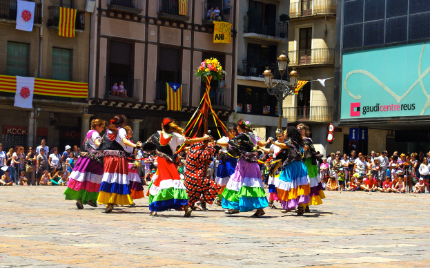 Eine Gruppe von Frauen in bunten Kleidern, die auf der Straße tanzen, umgeben von einer Menge Menschen, mit Gebäuden, Geländern, Balkonen, Flaggen, einem Laternenmast und einer Tafel mit Schrift im Hintergrund.